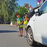 Zwei Schulkinder und eine Polizistin stehen hinter einem Auto und möchten die Straße überqueren.